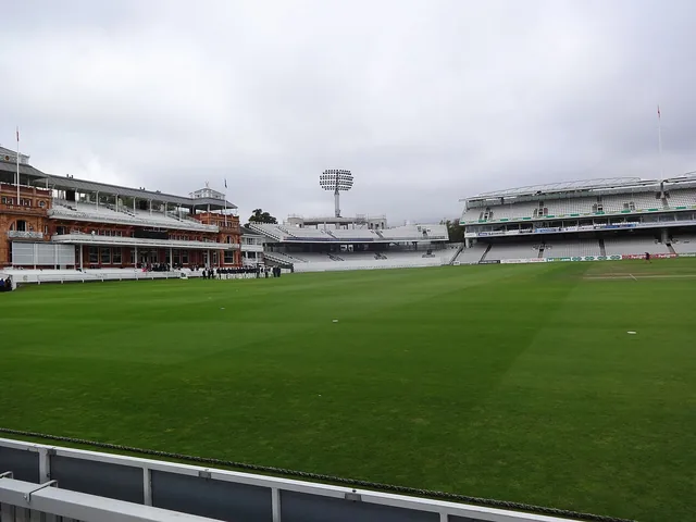 A grass cricket ground surrounded by stadium seating and a pavilion.