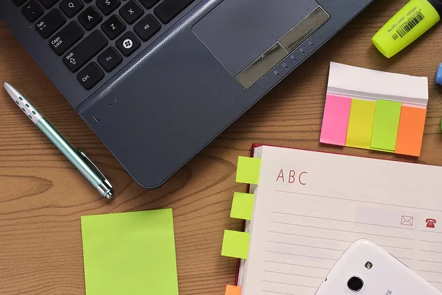 An open notebook, pen and page markers beside a laptop.
