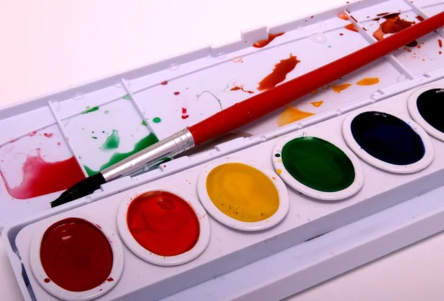 A paintbrush resting across a watercolour palette with wet colours in its pans.